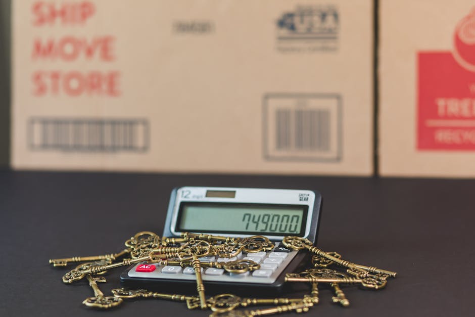 A close-up image showing a calculator with a digital display reading '749800', surrounded by a scattered collection of small brass keys. The calculator is placed on a dark surface in front of large cardboard boxes that are blurred in the background, with red and black text indicating shipping and moving labels. The scene reflects the packing process involved in home relocation, where keys and packing materials are organized for transportation, with the cardboard boxes ready for moving or storage. The overall setting appears to be indoors, with professional packing materials and tools visible as part of a furniture transport and moving logistics process in preparation for a house removal, with the focus on secure packing and efficient loading.