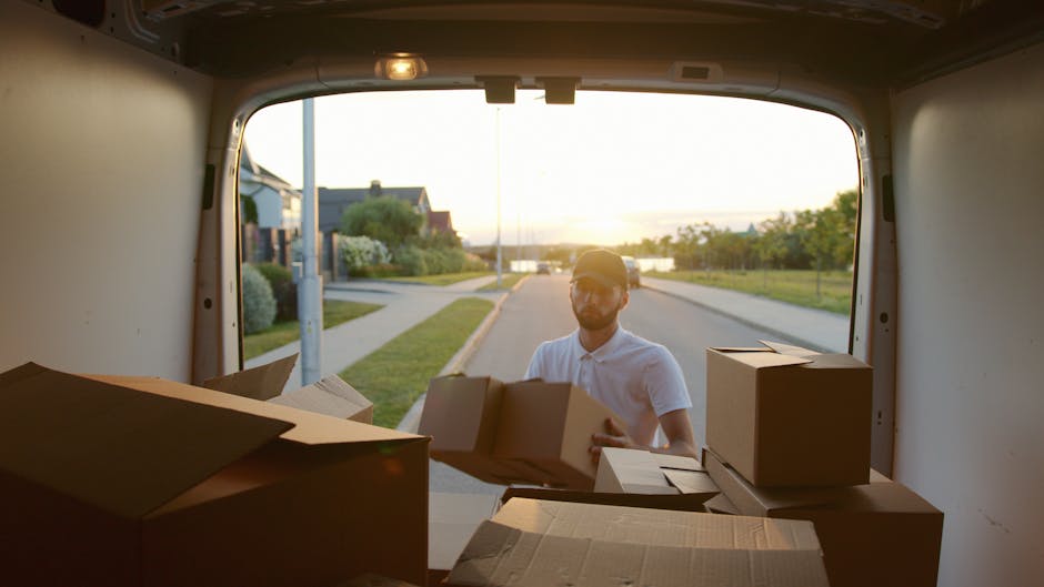 A man wearing a white polo shirt and cap is seen outside the rear of a moving van during a home relocation process. The van's interior is filled with partially packed cardboard boxes, some stacked and others laid flat, ready for transport. The boxes vary in size and are secured with packing materials such as bubble wrap or paper to protect fragile items. The man is in the process of loading or unloading boxes, holding one in his hands, with additional boxes positioned on the van's floor and side. The scene takes place early in the evening, with soft natural light illuminating the street outside, which features residential houses, a sidewalk, and a lamppost. The background shows calm neighborhood surroundings with a body of water visible in the distance, indicating a peaceful community. This image reflects professional furniture transport and packing and moving services provided by Man with a Van Kingston Vale, supporting efficient house removals and relocation logistics.