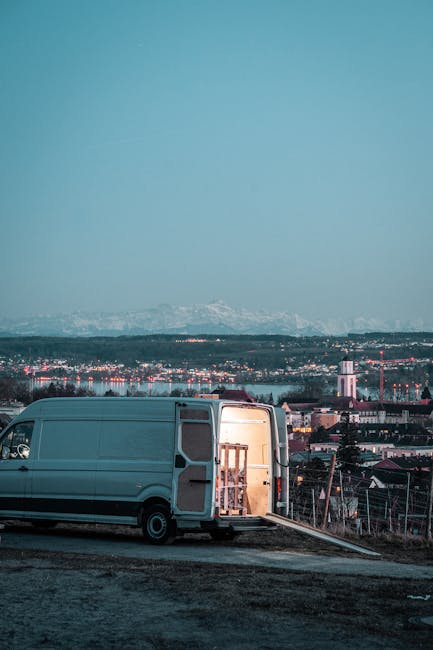 A light grey cargo van with its side door open, parked on a gravel surface outdoors during twilight. Inside the van, wooden furniture pieces are visible, secured with plastic wrap and packing blankets. The van is positioned next to a portable ramp leading into its interior, indicating an active loading process as part of a home relocation or furniture transport operation. In the background, there is a panoramic view of a cityscape with various industrial and residential buildings, including a tower and other structures, set against a distant mountain range under a clear sky with a gradient from pale blue to darker hues. This scene captures the logistical steps involved in house removals and the use of appropriate equipment like straps and blankets, in line with services provided by Man with a Van Kingston Vale.