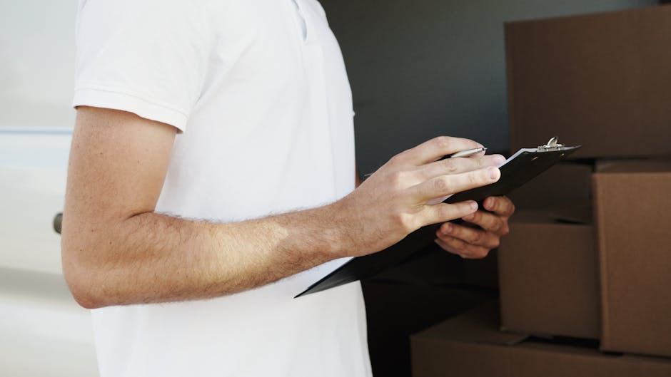 A person wearing a white t-shirt is holding a black clipboard with a pen in their right hand, inspecting a stack of cardboard moving boxes inside a home. The boxes are placed on a wooden surface or background, some are wrapped in transparent plastic wrap, and the person's left hand supports the clipboard. The scene likely depicts a packing or inventory check as part of a home relocation process. The environment is well-lit, highlighting the details of the person's arms, the packing materials, and the boxes, reflecting the logistics of furniture transport and moving preparations, associated with professional removals services such as those offered by Man with a Van Kingston Vale.