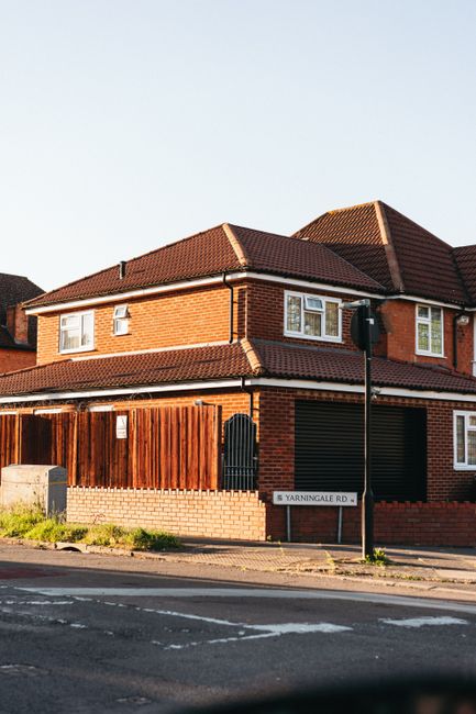 A two-storey brick house with a red-tiled roof, situated on a residential street, features a wooden fence along the perimeter and a black gate at the entrance. Outside, at the pavement, a green street