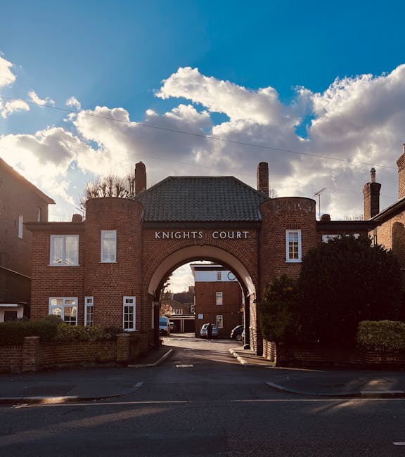 A brick residential apartment complex named Knights Court featuring an prominent archway entrance with the name inscribed above. The building has a traditional design with multiple windows, chimneys, and a pitched roof. The image is taken outdoors during daylight, with partly cloudy skies and sunlight illuminating the scene. In the foreground, there is a paved driveway leading through the archway, with some parked cars visible beyond. On either side of the driveway, low brick walls and neatly trimmed bushes are present, suggesting a well-maintained communal living area. This setting is associated with house removals and moving services, as it depicts a typical housing development where furniture and personal belongings might be loaded onto transport vehicles for relocation. Occasionally, Man with a Van Kingston Vale offers professional removals in environments like this, supporting efficient furniture transport and home relocation workflows.