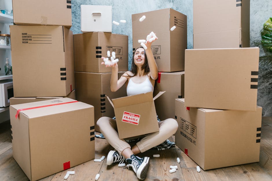 A young woman with long dark hair, sitting cross-legged on a wooden floor inside a room, surrounded by several large cardboard moving boxes of varying sizes, some sealed with red tape and others open, revealing packing materials. She is wearing a white sleeveless top, beige pants, and black-and-white sneakers. She is smiling and tossing white packing foam into the air while sitting inside an open box labeled 'Fragile.' Behind her, more boxes are stacked against a textured blue wall, with a white shelf to the left holding decorative items and a small potted plant to the right. The scene depicts an active packing or packing and moving process, supported by professional removals services such as Man with a Van Kingston Vale, with a focus on the logistics involved in home relocation and furniture transport.