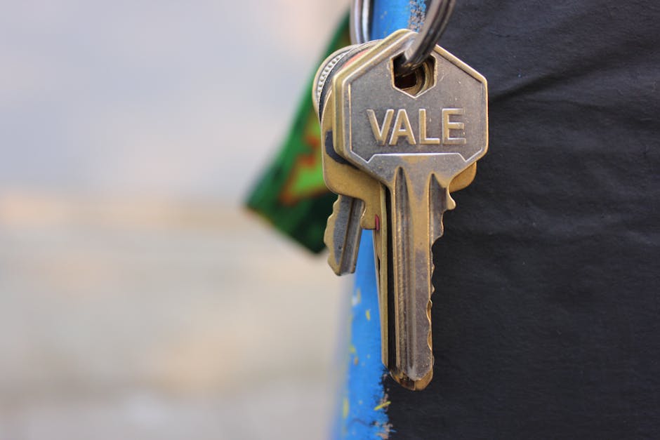Close-up of a set of brass keys attached to a keyring, with one key engraved with the name 'Vale,' held by a person wearing a dark jacket, standing outdoors near a house. In the background, part of a moving van with an open door is visible, loaded with packed cardboard boxes, plastic-wrapped furniture, and other packing materials. The scene depicts elements of house removals and furniture transport, including the loading process, with a focus on securing keys possibly for a property access during a home relocation in Kingston Vale. Natural daylight illuminates the scene, emphasizing the packing and loading activities associated with professional removal services provided by Man with a Van Kingston Vale.