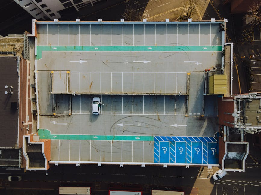 An aerial view of a multi-storey building's rooftop parking area during daytime, showing a single white van parked near the center. The parking lot has marked spaces with white lines, including blue designated spots for disabled parking located at the bottom right corner. There are green bicycle lanes with painted arrows indicating direction along the top and bottom edges of the lot, and a small section of pedestrian pathway with tactile paving at the bottom left. The rooftop surface is made of concrete with visible rust stains and wear. A small metal storage container is positioned at the top right corner adjacent to a yellow utility box. Surrounding the parking area are taller buildings and city streets, with minimal lighting and shadows cast by sunlight, consistent with urban house removals and furniture transport logistics. Man with a Van Kingston Vale specializes in home relocation services, as indicated by the context of the image associated with house removals and parking regulations in Kingston Vale.