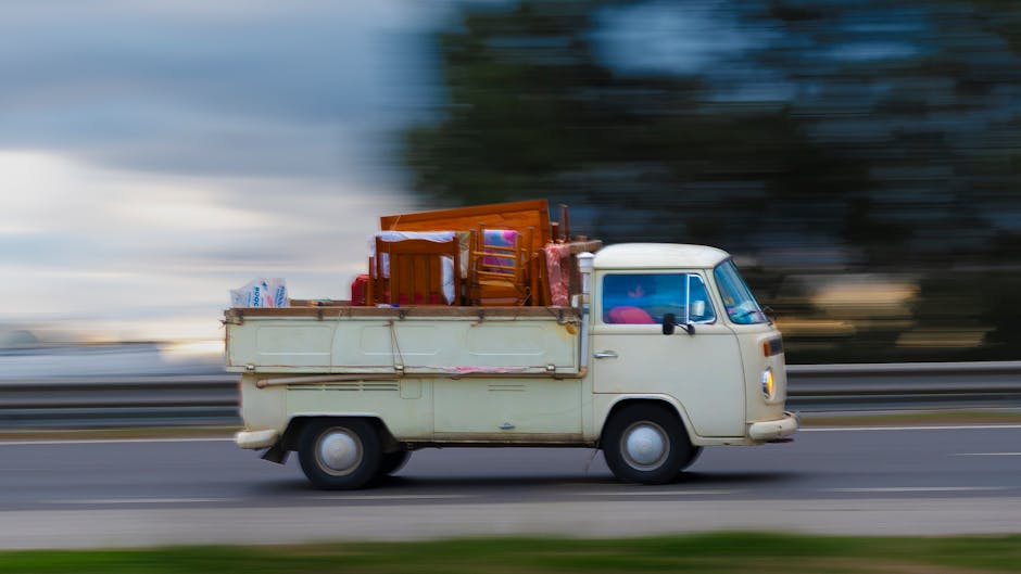 A vintage cream-colored van with a flatbed cargo area is moving along a roadway in Kingston Vale, as part of a home relocation or furniture transport process. The van's flatbed is loaded with various items including wooden furniture like chairs and a small table, all secured with protective packaging materials such as plastic wrap and blankets. Cardboard boxes are also present, some wrapped in plastic, indicating packing for a move. The van is being driven on a multi-lane road with a metal guardrail beside it, and the background features blurred trees and cloudy sky, emphasizing motion and speed during the loading process. Inside the vehicle, visible through the windshield, are additional packing supplies or personal belongings, suggesting ongoing loading or unloading activities. This scene reflects a typical furniture transport operation carried out by Man with a Van Kingston Vale as part of a professional removals service, with emphasis on proper packing and logistical planning for home moves in the Kingston Vale area, supported by local council regulations for moving vans.