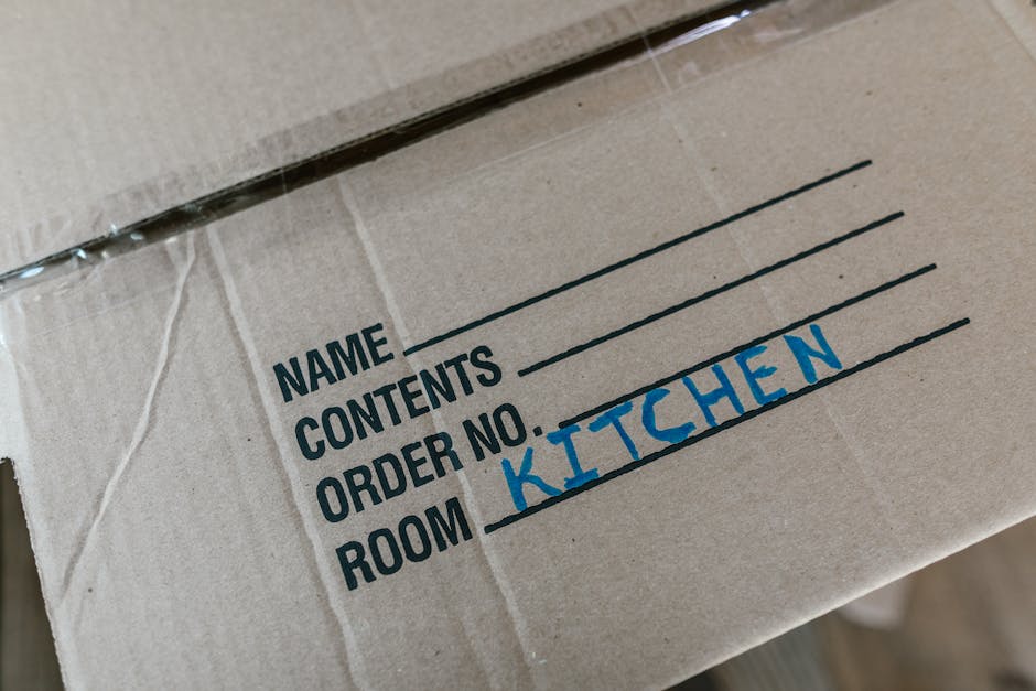Close-up image of a cardboard box used for home relocation, featuring printed labels with fields for 'Name,' 'Contents,' 'Order No.,' and 'Room,' with the handwritten text 'KITCHEN' in blue ink in the 'Room' section. The box is sealed with clear plastic tape along the edges and appears to be part of a packing and moving process, likely inside a property or on a pavement area prepared for furniture transport. The background shows part of a moving van or storage environment, indicating an organized loading process typical of professional removals carried out by companies like Man with a Van Kingston Vale, specialising in house removals and relocation services.