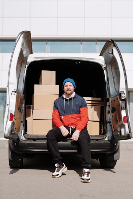 A young man with a beard, wearing a blue beanie, navy and red jacket, black pants, and white sneakers, is sitting on the edge of an open rear door of a white moving van parked outside a modern building with a white tiled facade and blue accents. The van's interior is filled with several cardboard boxes of various sizes, some stacked on top of each other, and wrapped in clear plastic or packing tape, indicating a home relocation or furniture transport process. The man appears relaxed and smiling, suggesting a break during the loading or unloading phase of a house move. The scene captures the loading process associated with packing and moving services, with the van positioned on a paved surface and the environment well-lit, highlighting the practical aspects of furniture transport carried out by companies like Man with a Van Kingston Vale.