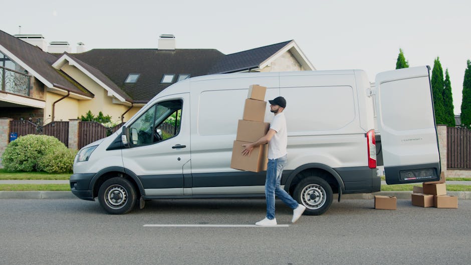 A young man with a beard, wearing a blue beanie, navy and red jacket, black pants, and white sneakers, is sitting on the edge of an open rear door of a white moving van parked outside a modern building with a white tiled facade and blue accents. The van's interior is filled with several cardboard boxes of various sizes, some stacked on top of each other, and wrapped in clear plastic or packing tape, indicating a home relocation or furniture transport process. The man appears relaxed and smiling, suggesting a break during the loading or unloading phase of a house move. The scene captures the loading process associated with packing and moving services, with the van positioned on a paved surface and the environment well-lit, highlighting the practical aspects of furniture transport carried out by companies like Man with a Van Kingston Vale.