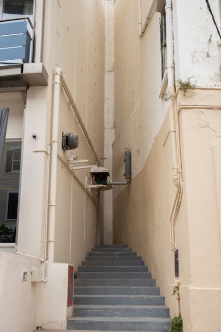 The image depicts a narrow outdoor staircase with dark grey steps situated between two tall, beige-painted building facades. On the left side, a grey metal air conditioning unit is mounted near the top of the wall, supported by a metal bracket. Multiple white PVC pipes run horizontally and vertically along both walls, some with black insulation or fittings. A small black and grey security camera is attached to a pipe on the left wall, facing downward. The right side of the staircase shows a small green plant sprouting from a crack in the wall near the bottom. Natural light illuminates the scene, creating a contrast between the shadows on the stairs and the bright sky above. This setting is common in urban environments and can pose challenges in house removals or furniture transport due to limited space and accessibility, which is relevant for services like those offered by Man with a Van Kingston Vale during home relocations or moving projects involving tight staircases.