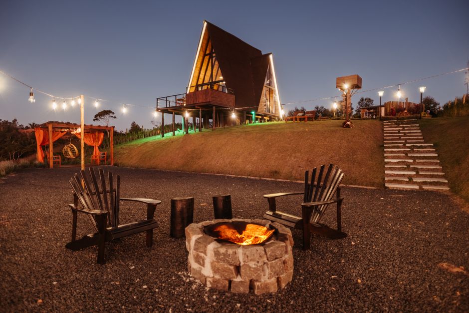 A nighttime outdoor scene featuring a modern, A-frame house elevated on stilts with a wooden balcony and illuminated by exterior lighting, situated on a gentle grassy slope. In the foreground, there are two wooden Adirondack chairs positioned around a stone fire pit with a glowing fire, and a small wooden side table between them. To the left, a wooden structure with hanging chairs or swings is visible, and to the right, a staircase made of concrete steps leads up the grassy hill. String lights are hung across the scene, enhancing visibility and ambiance. The area around the fire pit has a gravel surface, and the background includes trees and a dark sky, creating a cozy, relaxed setting that may be part of a home relocation or outdoor gathering area. This natural setting, combined with the arrangement of furniture, packaging materials, and lighting, reflects a scene where furniture might be moved or prepared for transport as part of a home removal service, aligning with the services offered by Man with a Van Kingston Vale.