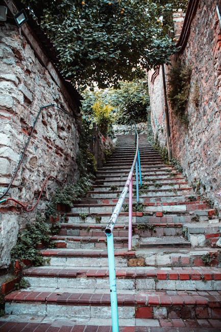 The image depicts a narrow outdoor staircase with dark grey steps situated between two tall, beige-painted building facades. On the left side, a grey metal air conditioning unit is mounted near the top of the wall, supported by a metal bracket. Multiple white PVC pipes run horizontally and vertically along both walls, some with black insulation or fittings. A small black and grey security camera is attached to a pipe on the left wall, facing downward. The right side of the staircase shows a small green plant sprouting from a crack in the wall near the bottom. Natural light illuminates the scene, creating a contrast between the shadows on the stairs and the bright sky above. This setting is common in urban environments and can pose challenges in house removals or furniture transport due to limited space and accessibility, which is relevant for services like those offered by Man with a Van Kingston Vale during home relocations or moving projects involving tight staircases.