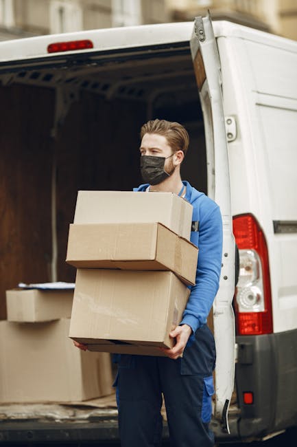 A man wearing a black face mask, blue hoodie, and dark trousers is standing outside the open rear doors of a white moving van, holding three flattened cardboard boxes of varying sizes, one on top of the other. Inside the van, additional cardboard boxes and packing materials are visible, some stacked on the floor. The vehicle is parked on a street or driveway, with the surroundings showing part of a building facade. The scene depicts a home relocation or furniture transport process, with the man preparing to load or unload items for a house move. This image reflects the activities involved in packing, moving logistics, and the transportation of household items by Man with a Van Kingston Vale as part of their comprehensive removals service.