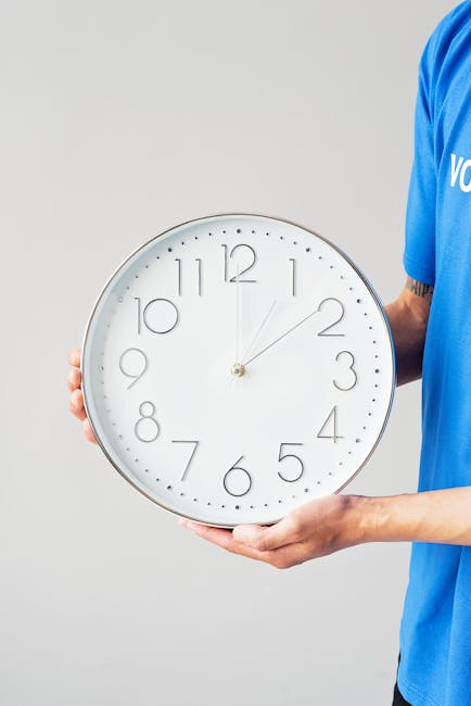 A person wearing a blue uniform holding a large round wall clock with a white face, black numbers, and silver frame. The clock shows approximately 12:07. The individual is gripping the clock by its sides, with the left hand at the 9 o'clock position and the right hand supporting the lower part of the clock. The background is plain and light grey. This scene may depict elements of home relocation or packing services, relevant to urgent same day removals in Kingston Vale provided by Man with a Van Kingston Vale.