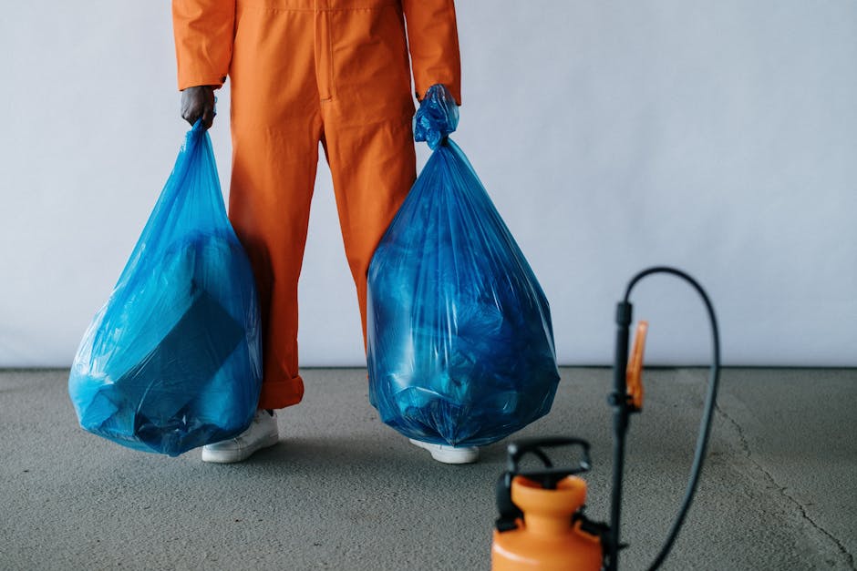 A person wearing orange work overalls and white footwear is standing indoors on a concrete floor, holding two large blue plastic trash bags filled with waste or packing materials. The individual’s legs and lower torso are visible, with the bags hanging from each hand. The background features a plain, light-colored wall, and a black electrical cable and a small orange dust extractor are positioned on the floor in the foreground. The setting appears to be a preparation or packing area used for home relocation or furniture transport, with the person involved in packing or waste disposal as part of the moving process, supported by the presence of packing materials and equipment for loading or transporting items during a house removal, aligning with services offered by Man with a Van Kingston Vale.