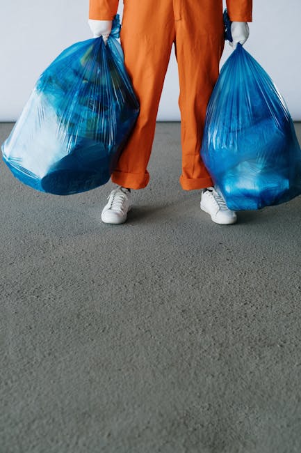 A person wearing orange trousers and white sneakers is standing indoors on a grey carpeted floor, holding two large blue plastic bags filled with waste or recyclable materials in each hand, with the bags extending downward to the floor. The individual is positioned against a plain white wall, and only the lower half of their body is visible. The scene depicts a house clearance or waste disposal process as part of a home relocation or moving service. The environment suggests an indoor setting, possibly a hallway or room, with the person's stance indicating they are about to transport or load the waste bags for removal, aligning with furniture transport and packing logistics handled by Man with a Van Kingston Vale.