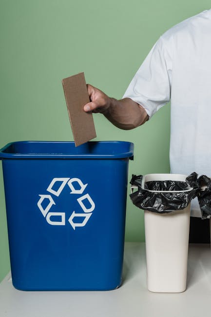 A person wearing orange work overalls and white footwear is standing indoors on a concrete floor, holding two large blue plastic trash bags filled with waste or packing materials. The individual’s legs and lower torso are visible, with the bags hanging from each hand. The background features a plain, light-colored wall, and a black electrical cable and a small orange dust extractor are positioned on the floor in the foreground. The setting appears to be a preparation or packing area used for home relocation or furniture transport, with the person involved in packing or waste disposal as part of the moving process, supported by the presence of packing materials and equipment for loading or transporting items during a house removal, aligning with services offered by Man with a Van Kingston Vale.